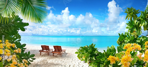 Beautiful tropical beach with white sand and two sun loungers on background of turquoise ocean and blue sky with clouds. Frame of palm leaves and flowers. Perfect landscape for relaxing vacation.