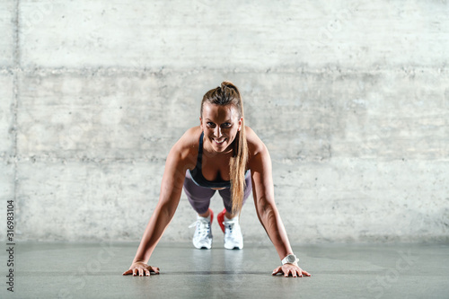 Young attractive smiling positive caucasian sportswoman in sportswear and with ponytail doing push ups indoors and looking at camera. Healthy lifestyle concept.