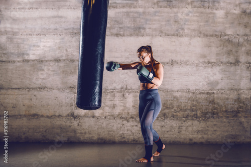 Full length of young dedicated caucasian muscular female boxer in sportswear with ponytail and boxing gloves punching boxing bag while standing in the gym. © dusanpetkovic1