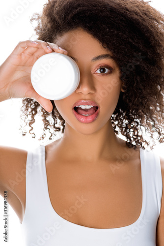 surprised african american girl holding plastic container with face cream, isolated on white