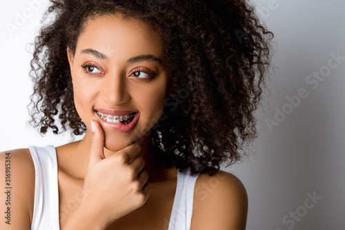 thoughtful smiling african american girl with dental braces touching lip, isolated on grey
