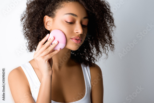 curly african american girl using silicone cleansing facial brush, isolated on grey