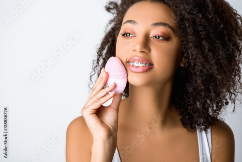 smiling african american girl with dental braces using silicone cleansing facial brush, isolated on grey