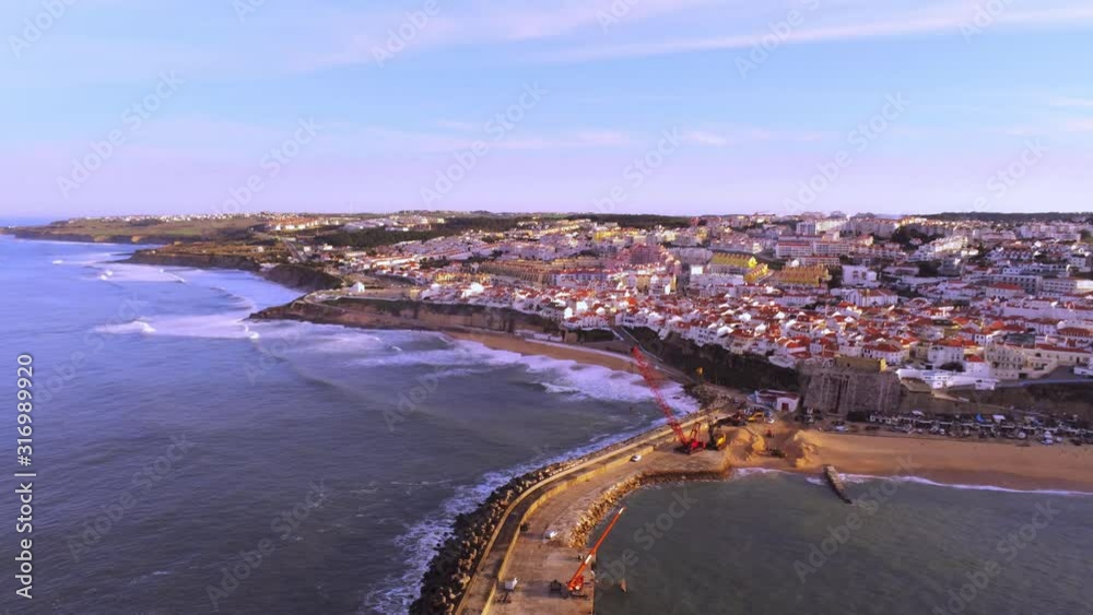 Elevated Island Full Of Beautiful Houses With Tower Cranes Working By The Shore In Ericeira, Portugal On A Hot Sunny Weather - Aerial Shot Elevated Island Full Of Beautiful Houses With Tower Cranes Working By The Shore In Ericeira, Portugal On A Hot Sunny Weather - Aerial Shot