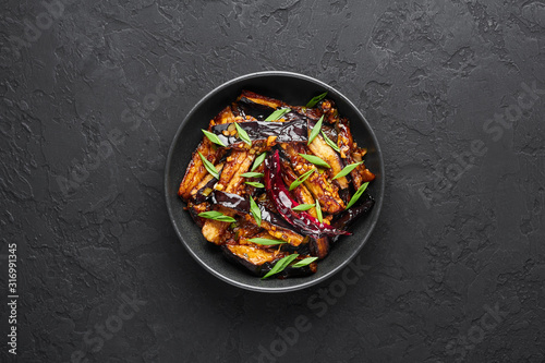 Sichuan Eggplant Stir Fry in black bowl at dark slate background. Eggplant Stir Fry is chinese cuisine dish with deep fried eggplant, chilli pepper, different sauces. Copy space. Top view