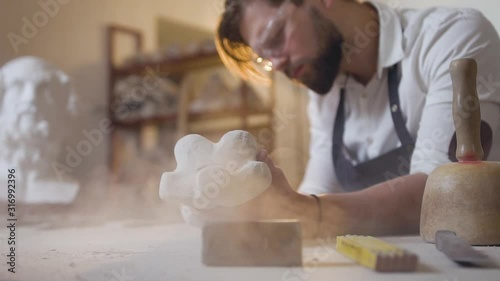 Neat careful likable bearded sculptor working in his studio with polishing tool and handmade figure from chalk-stone photo