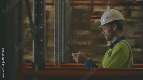 Male Industrial Worker in the Hard Hat Uses Mobile Phone While Walking Through Warehouse photo