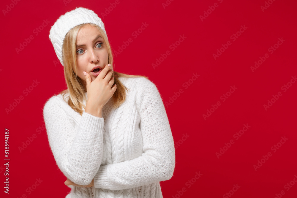 Shocked young woman in white sweater hat isolated on red wall background, studio portrait. Healthy fashion lifestyle, cold season concept. Mock up copy space. Looking aside, put hand prop up on chin. Shocked young woman in white sweater hat isolated on red wall background, studio portrait. Healthy fashion lifestyle, cold season concept. Mock up copy space. Looking aside, put hand prop up on chin.
