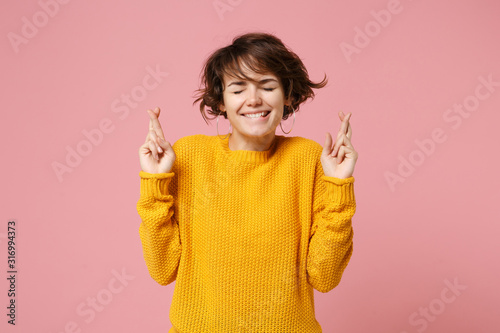 Young brunette woman in yellow sweater posing isolated on pink background. People lifestyle concept. Mock up copy space. Waiting for special moment, keeping fingers crossed, eyes closed, making wish.