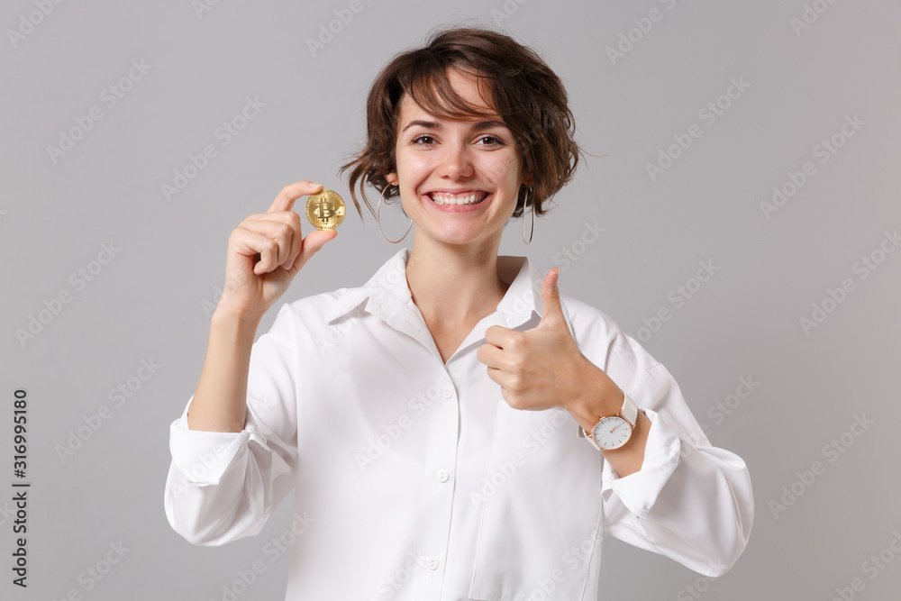 Smiling young business woman in white shirt posing isolated on grey wall background. Achievement career wealth business concept. Mock up copy space. Showing thumb up, hold bitcoin, future currency. Smiling young business woman in white shirt posing isolated on grey wall background. Achievement career wealth business concept. Mock up copy space. Showing thumb up, hold bitcoin, future currency.