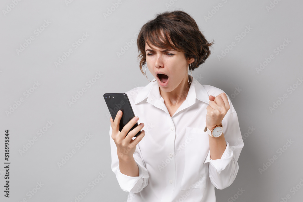 Angry irritated young business woman in white shirt posing isolated on grey background studio portrait. Achievement career wealth business concept. Mock up copy space. Holding mobile phone, swearing. Angry irritated young business woman in white shirt posing isolated on grey background studio portrait. Achievement career wealth business concept. Mock up copy space. Holding mobile phone, swearing.