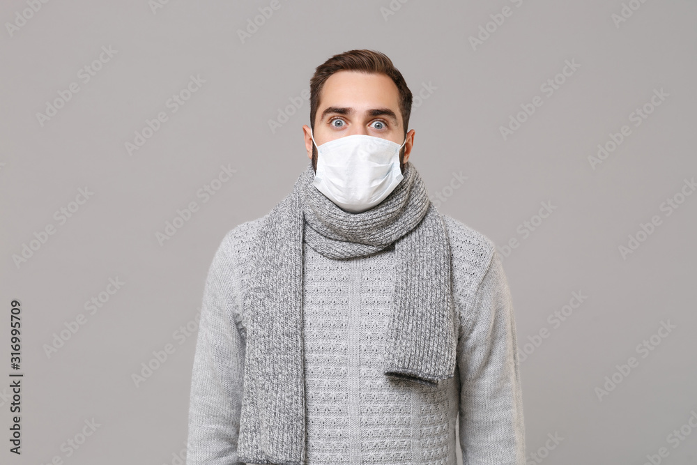 Shocked young man in gray sweater, scarf isolated on grey background studio portrait. Healthy lifestyle, ill sick disease treatment, cold season concept. Mock up copy space. Wearing sterile face mask. Shocked young man in gray sweater, scarf isolated on grey background studio portrait. Healthy lifestyle, ill sick disease treatment, cold season concept. Mock up copy space. Wearing sterile face mask.