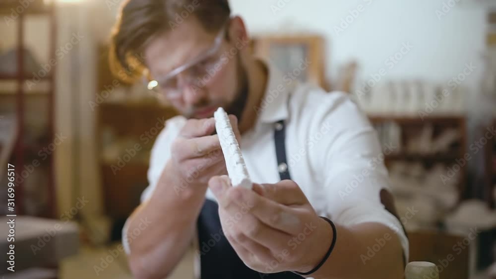 Good-looking careful young bearded master with fashion haircut wearing special glasses checking the detail to the sculpture Good-looking careful young bearded master with fashion haircut wearing special glasses checking the detail to the sculpture