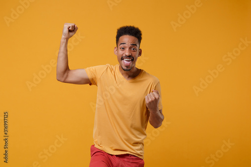 Excited young african american guy in casual t-shirt posing isolated on yellow orange wall background, studio portrait. People emotions lifestyle concept. Mock up copy space. Doing winner gesture.