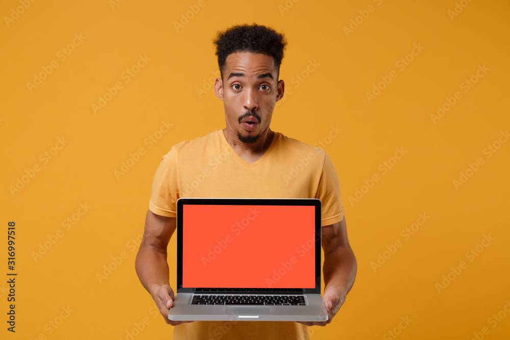 Shocked young african american guy in casual t-shirt posing isolated on yellow orange wall background. People lifestyle concept. Mock up copy space. Hold laptop pc computer with blank empty screen. Shocked young african american guy in casual t-shirt posing isolated on yellow orange wall background. People lifestyle concept. Mock up copy space. Hold laptop pc computer with blank empty screen.