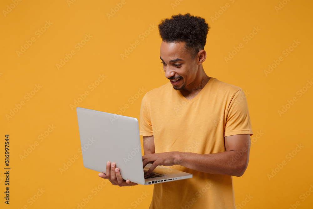 Smiling young african american guy in casual t-shirt posing isolated on yellow orange wall background, studio portrait. People lifestyle concept. Mock up copy space. Hold in hands laptop pc computer. Smiling young african american guy in casual t-shirt posing isolated on yellow orange wall background, studio portrait. People lifestyle concept. Mock up copy space. Hold in hands laptop pc computer.