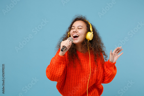Funny young african american girl in orange knitted clothes isolated on pastel blue background. People lifestyle concept. Mock up copy space. Listen music with headphones, sing song in microphone.