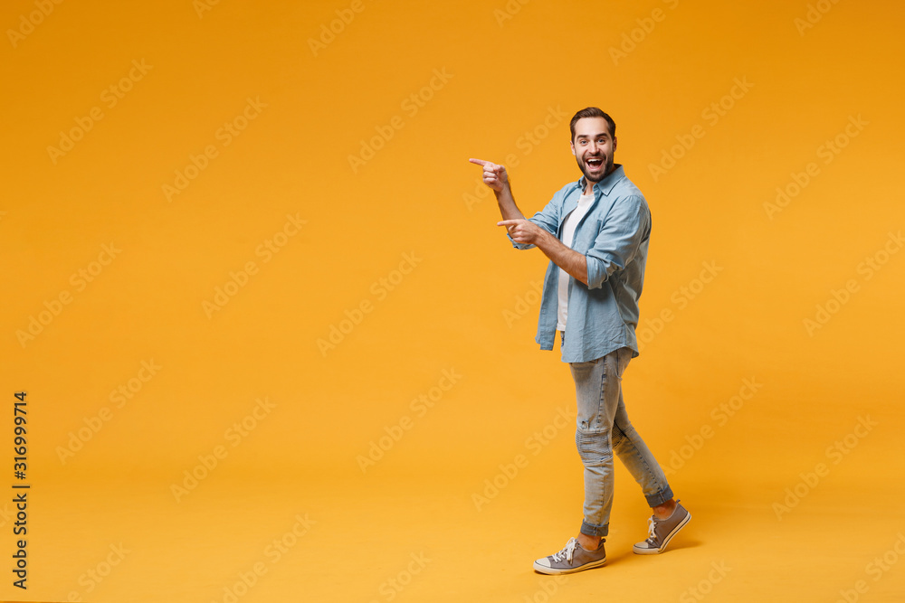 Excited young bearded man in casual blue shirt posing isolated on yellow orange background studio portrait. People sincere emotions lifestyle concept. Mock up copy space. Pointing index fingers aside. - obrazy, fototapety, plakaty 