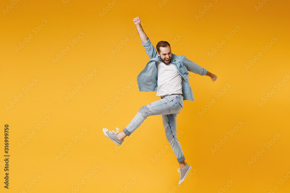 Funny young bearded man in casual blue shirt posing isolated on yellow orange wall background studio portrait. People sincere emotions lifestyle concept. Mock up copy space. Jumping, rising hands up.