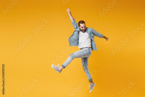Funny young bearded man in casual blue shirt posing isolated on yellow orange wall background studio portrait. People sincere emotions lifestyle concept. Mock up copy space. Jumping  rising hands up.