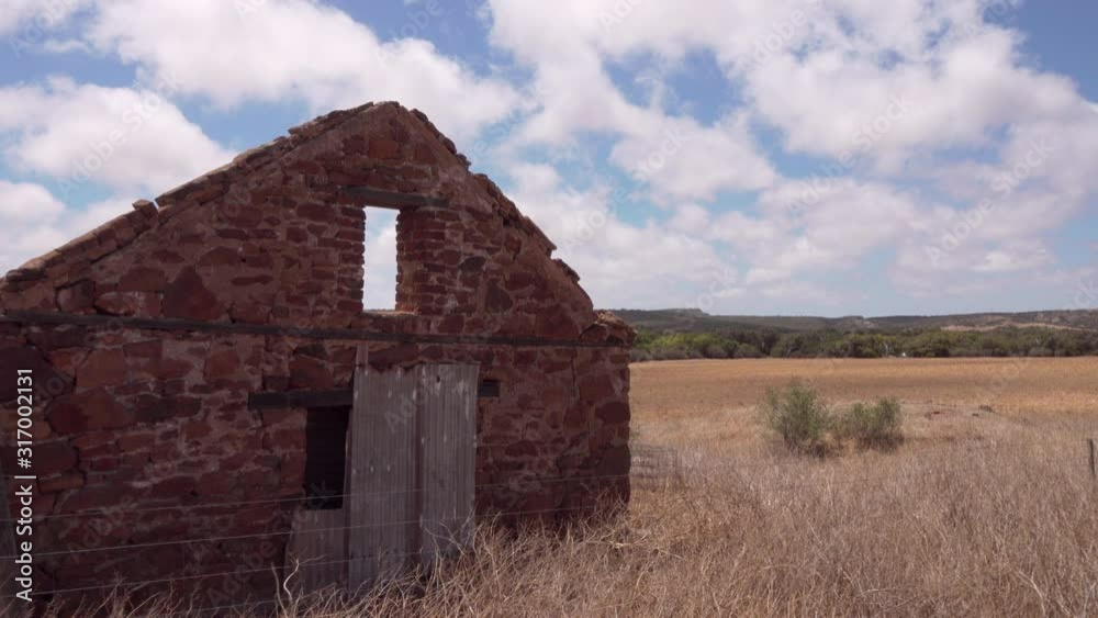 Zoom shot approaching ruins of an old stone house set in a large dry paddock covered in long brown grass. with low hills in background and patchy clouds in sky Zoom shot approaching ruins of an old stone house set in a large dry paddock covered in long brown grass. with low hills in background and patchy clouds in sky