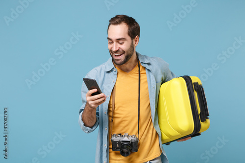 Cheerful traveler tourist man in casual clothes with photo camera isolated on blue background. Male passenger traveling abroad on weekends. Air flight journey concept Hold suitcase using mobile phone.