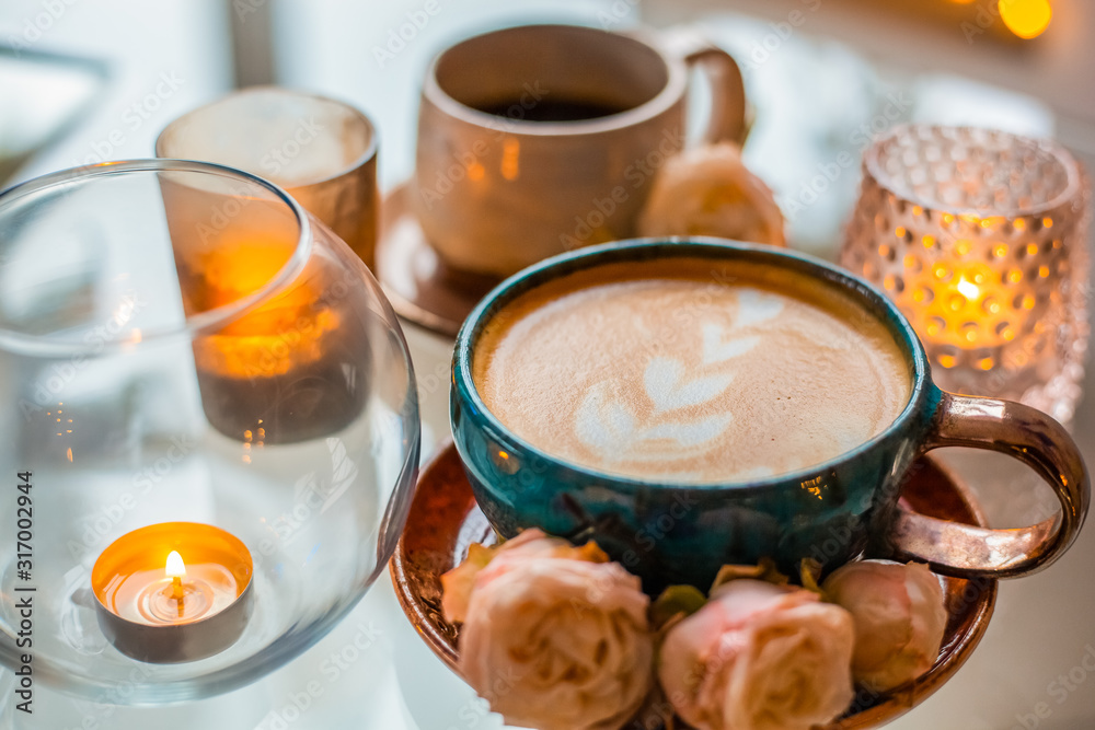 A cup of coffee on a saucer is decorated with roses next to candles in beautiful candlesticks on a glass table A cup of coffee on a saucer is decorated with roses next to candles in beautiful candlesticks on a glass table