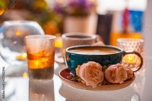 A cup of coffee on a saucer is decorated with roses next to candles in beautiful candlesticks on a glass table