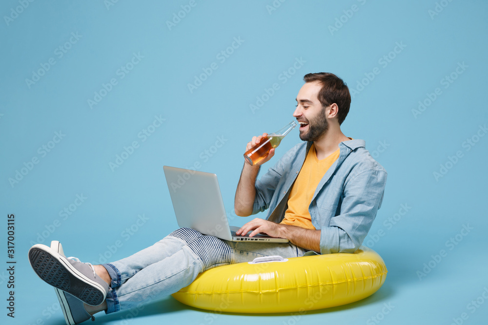 Cheerful traveler tourist man in yellow clothes isolated on blue background. Passenger traveling abroad on weekend. Air flight journey concept. Sit in inflatable ring work on laptop, hold beer bottle.