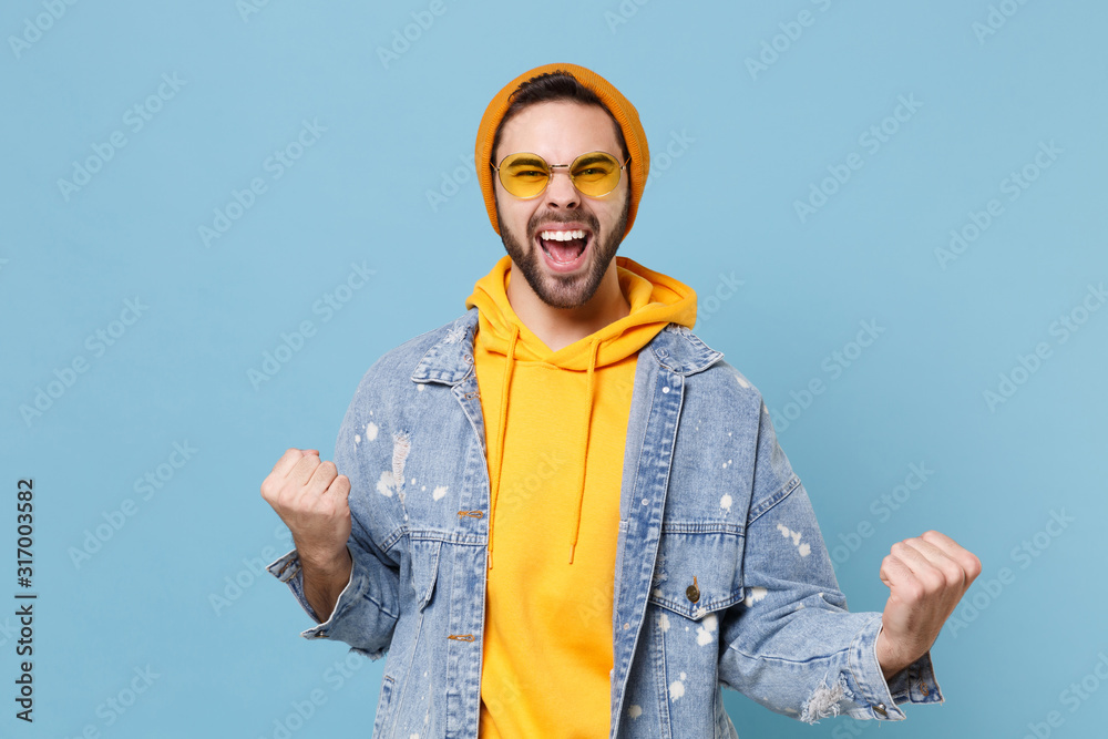 Happy young hipster guy in fashion jeans denim clothes posing isolated on pastel blue background studio portrait. People sincere emotions lifestyle concept. Mock up copy space. Doing winner gesture.