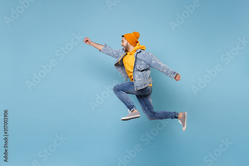 Side view of young hipster guy in fashion jeans denim clothes posing isolated on pastel blue background. People lifestyle concept. Mock up copy space. Jumping with outstretched hand like Superman.