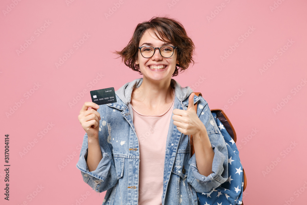 Smiling young woman student in denim clothes glasses backpack posing isolated on pastel pink background. Education in high school university college concept. Showing thumb up, hold credit bank card. Smiling young woman student in denim clothes glasses backpack posing isolated on pastel pink background. Education in high school university college concept. Showing thumb up, hold credit bank card.