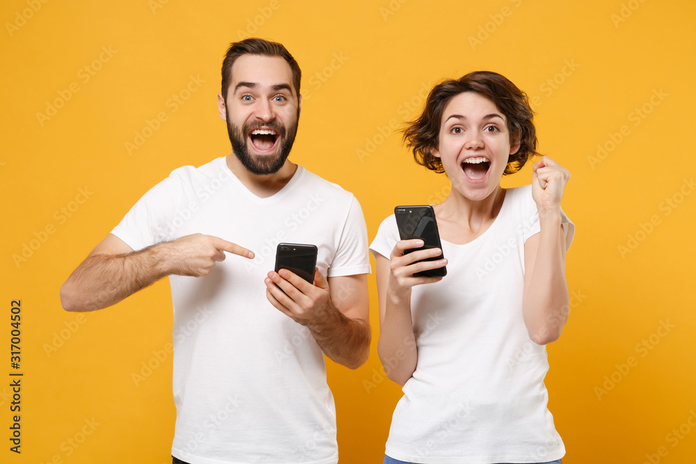 Excited couple friends bearded guy girl in white t-shirts isolated on yellow orange background. People lifestyle concept. Mock up copy space. Point index fingers on mobile phone, doing winner gesture.