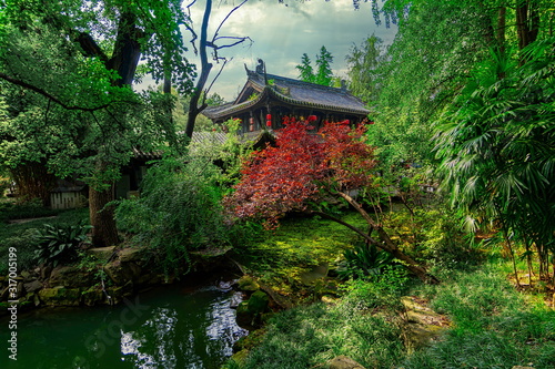 Small temple hidden in the middle of the vegetation. The green of the leaves contrasts with the red of the tree. Wuhou Temple, Chengdu city, China