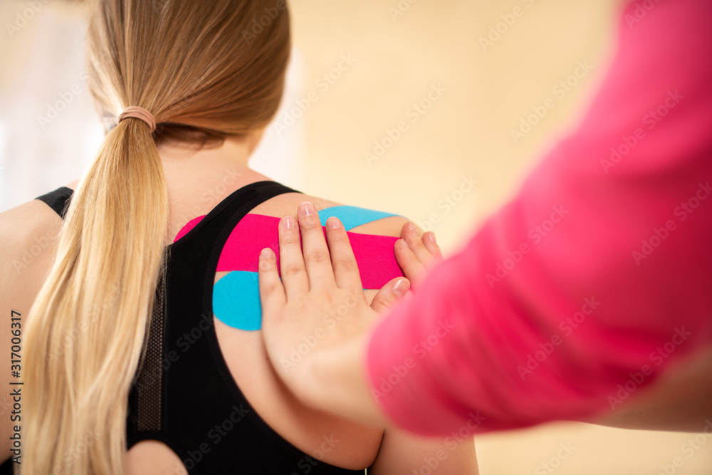 Physical therapist applying kinesio tape on female patient's shoulder. Close up rear view. Kinesiology, physical therapy, rehabilitation concept. Physical therapist applying kinesio tape on female patient's shoulder. Close up rear view. Kinesiology, physical therapy, rehabilitation concept.