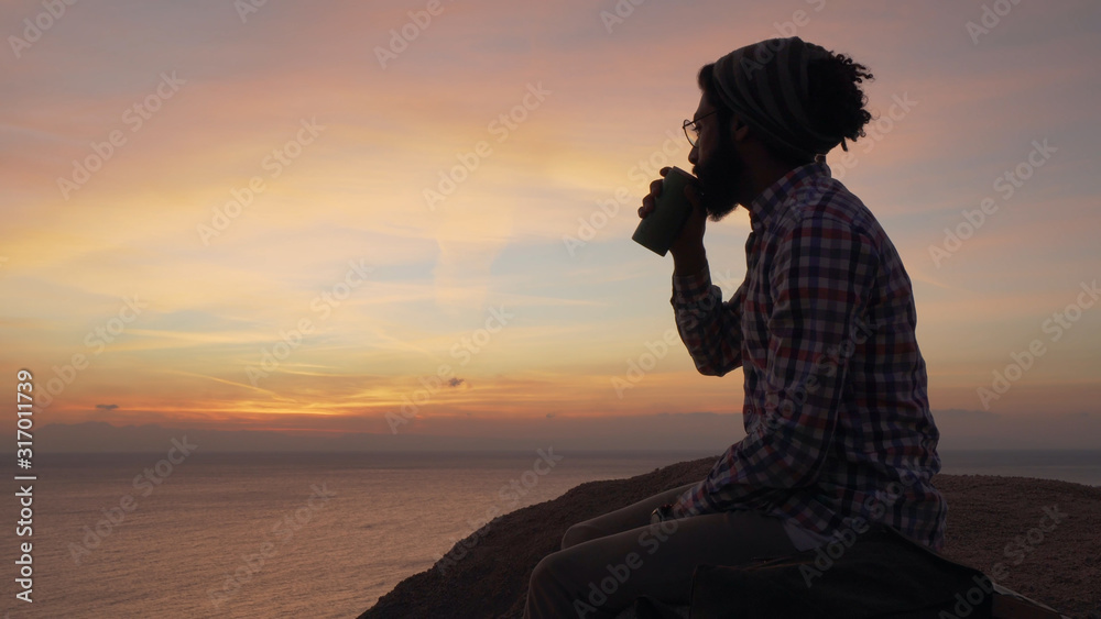 silhouette of male sitting on a cliff and drinking coffee at sunrise