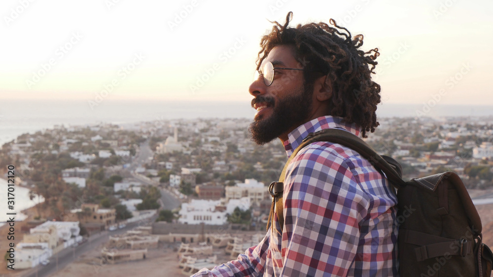 smiling adult traveler with dreadlocks sitting on a cliff at sunrise smiling adult traveler with dreadlocks sitting on a cliff at sunrise