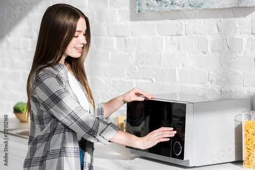 smiling and attractive woman in shirt using microwave in kitchen