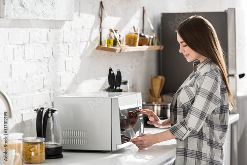 side view of smiling woman in shirt using microwave in kitchen photo