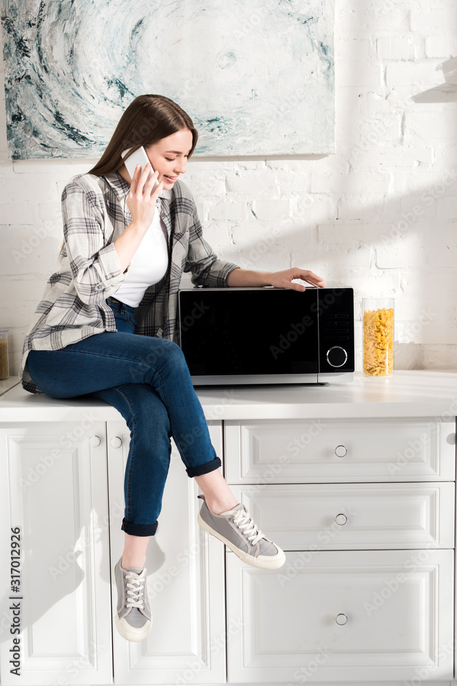 smiling woman talking on smartphone and looking at microwave in kitchen smiling woman talking on smartphone and looking at microwave in kitchen