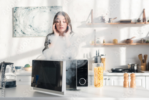 Obraz na plátně shocked and attractive woman looking at broken microwave in kitchen