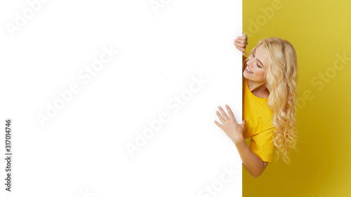 Young happy woman peeks out from behind a white banner on a yellow background. Point to an empty blank on a form, a copy space for text. Horizontal shot