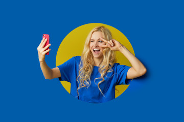Image of young happy woman standing over a blue background. Girl peep out of a hole in the wall. Looking to the side to take a selfie on the mobile phone.