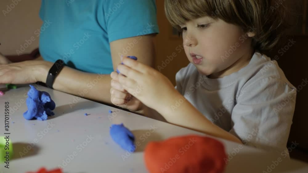 Child in Process of Modeling of Plasticine at Table in Nursery Room. Child Kid Boy and Mother Playing Colorful Clay Toy at Playroom of Home. Young Woman and Son Playing With Colorful Clay in Nursery. Child in Process of Modeling of Plasticine at Table in Nursery Room. Child Kid Boy and Mother Playing Colorful Clay Toy at Playroom of Home. Young Woman and Son Playing With Colorful Clay in Nursery.