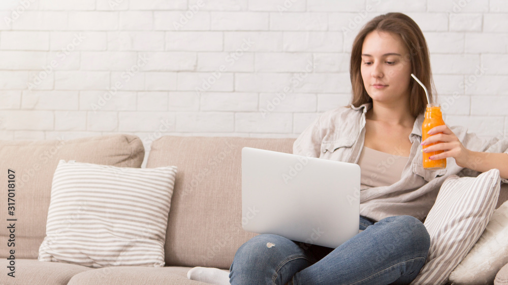 Woman drinking homemade fruity detox smoothie sitting on sofa and using laptop