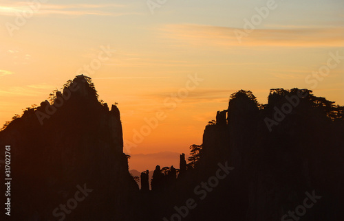 Huangshan Mountain in Anhui Province, China. Sunrise over Huangshan showing a detailed silhouette of the mountain ridge against an orange sky. Sunrise near the summit of Huangshan Mountain, China.