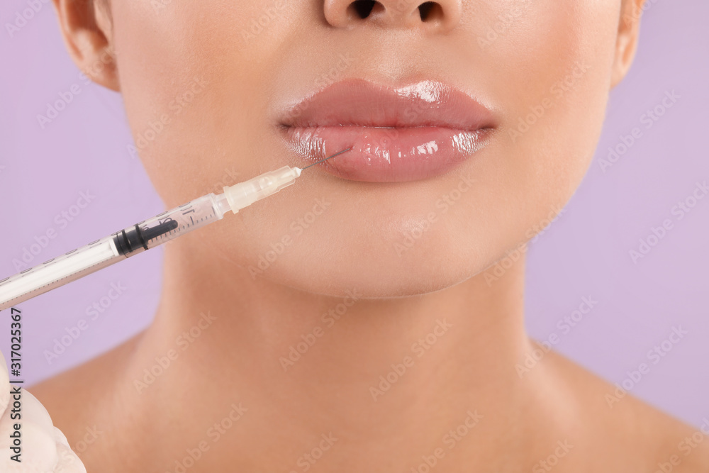 Young woman getting lips injection on lilac background, closeup