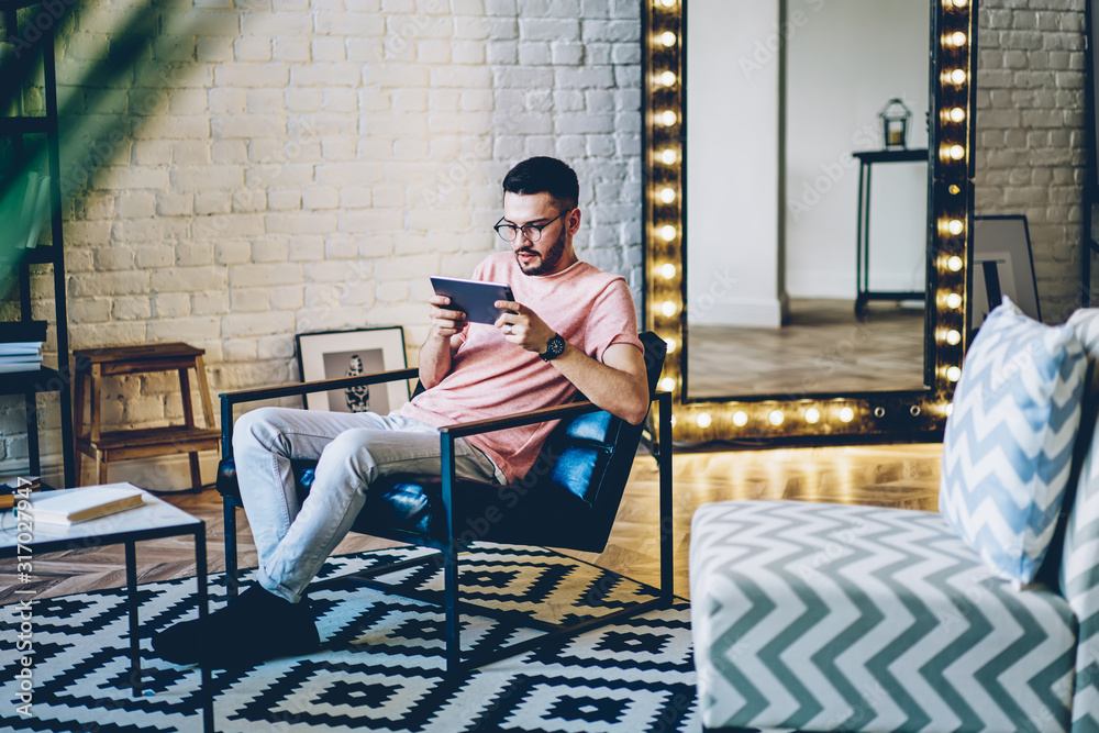 Fototapeta premium Young male blogger resting at own apartment with stylish comfortable interior and using modern touch pad for watching video online on web page, hipster guy relaxing at home with new digital tablet