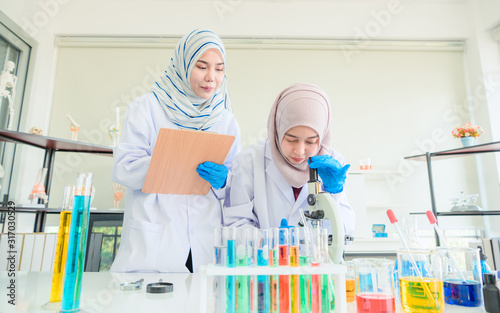 Beautiful female muslim chemists in hijab with arms crossed and clipboard looking at camera in laboratory