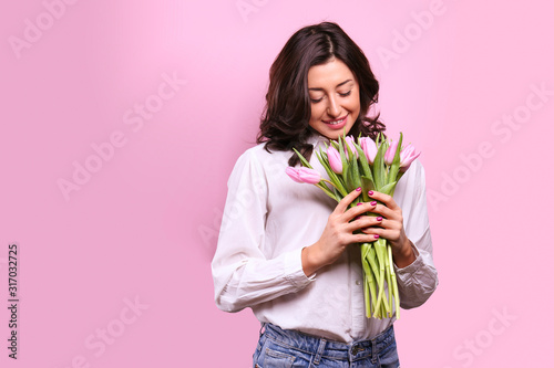 Studio portrait of gorgeous young brunette woman with long wavy hair wearing white loose cotton shirt, holding bouquet of tulip flowers. Pink isolated background, copy space, close up. #317032725